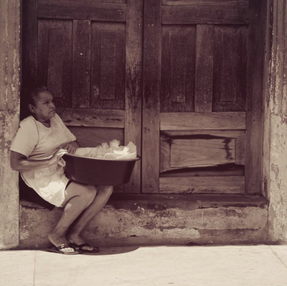 Sepia photo of elderly Hondurian woman sat in wooden doorway with basket of vegetables
